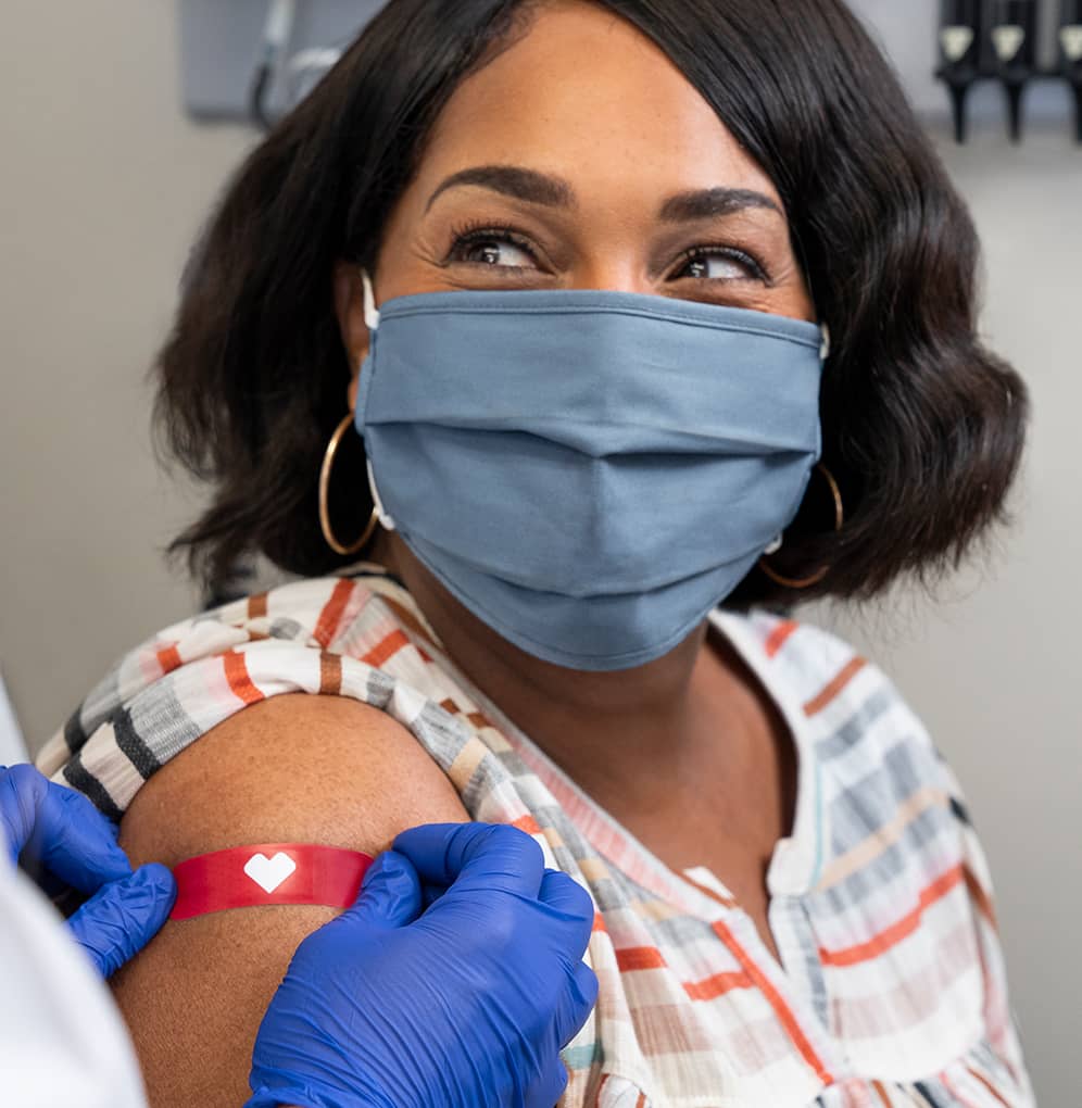 Learn about COVID-19 vaccine, showing a woman in a face covering as someone puts  a bandage on her arm, post-vaccination