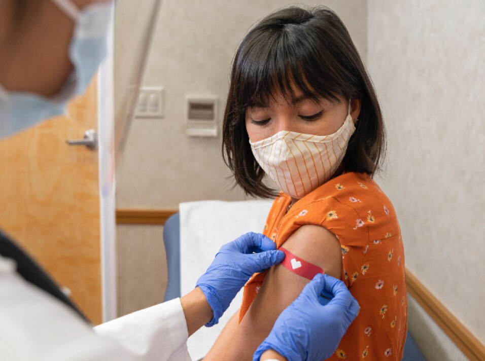 woman wearing a face covering, looking down at the bandage being applied to her upper arm after having been vaccinated