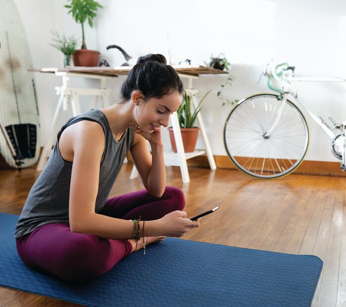 young woman seated on a yoga mat, looking down at her cell phone screen