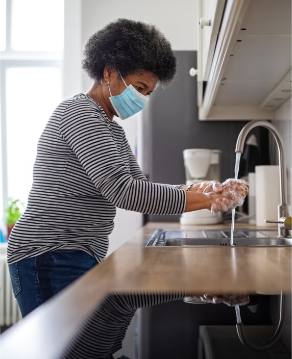 An image of woman in face covering washing her hands in a kitchen