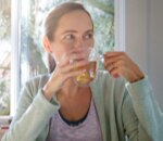 An adult woman sitting at a kitchen table sipping a mug of tea, deep in thought