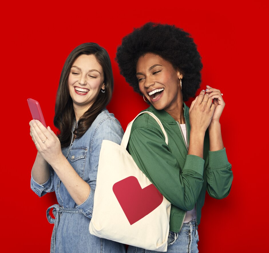 Two young-adult women standing back to back, smiling and looking at the CVS app on a mobile phone.