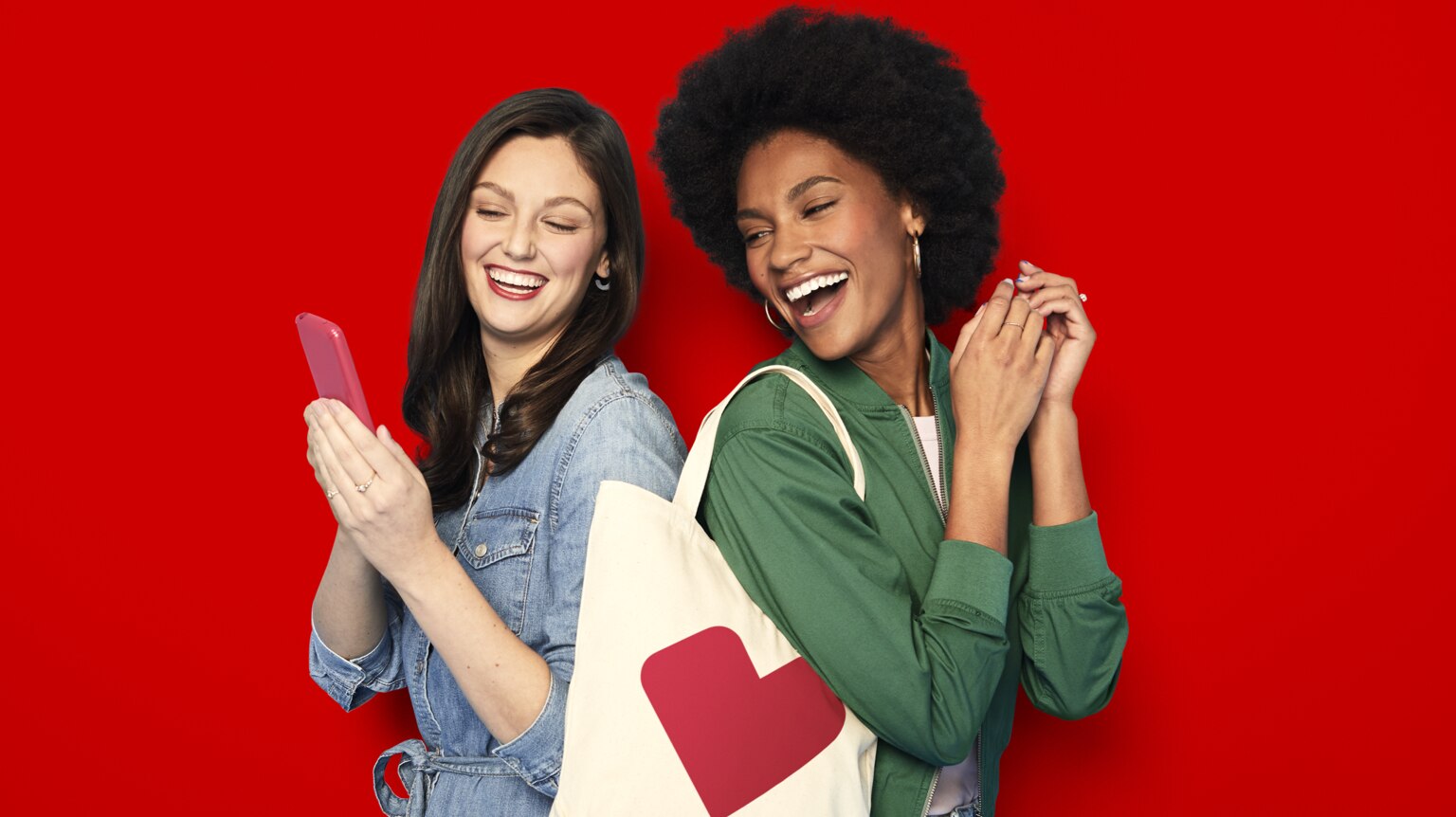 Two young-adult women standing back to back, smiling and looking at the CVS app on a mobile phone.