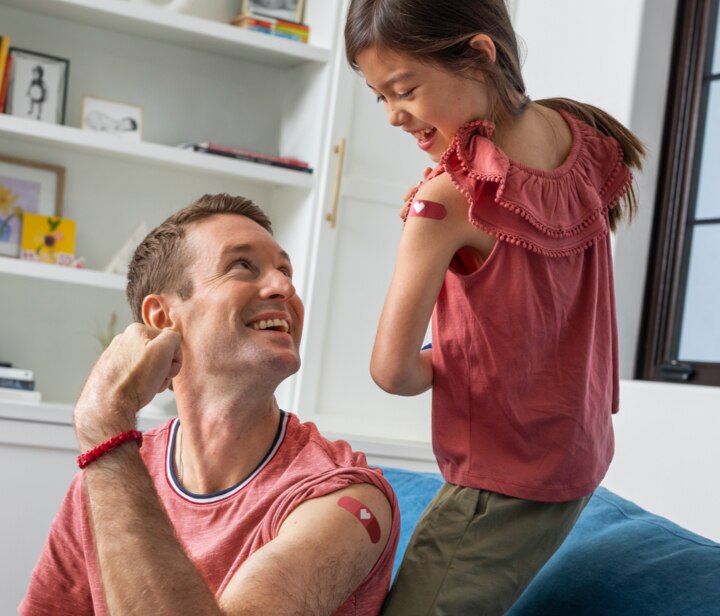 Smiling father and young daughter showing off matching CVS bandages on their arms after receiving vaccinations