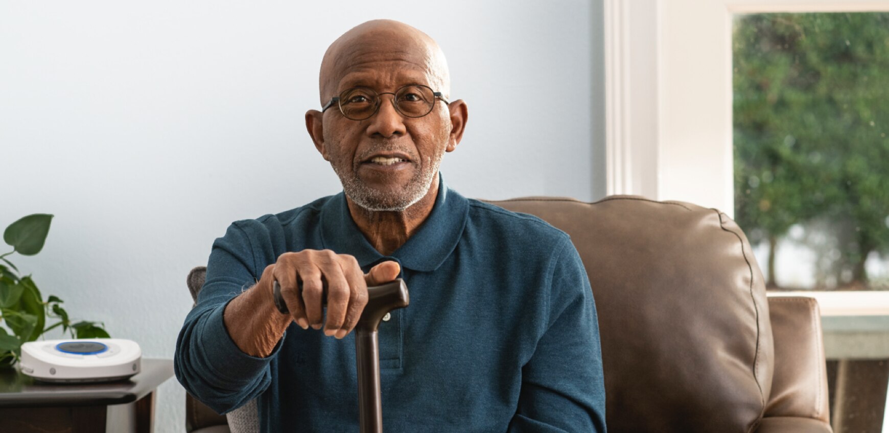 Smart Hub out of focus on living room side table. Elder gentleman sitting on couch holding walking cane.