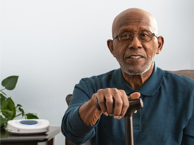 Smart Hub out of focus on living room side table. Elder gentleman sitting on couch holding walking cane.