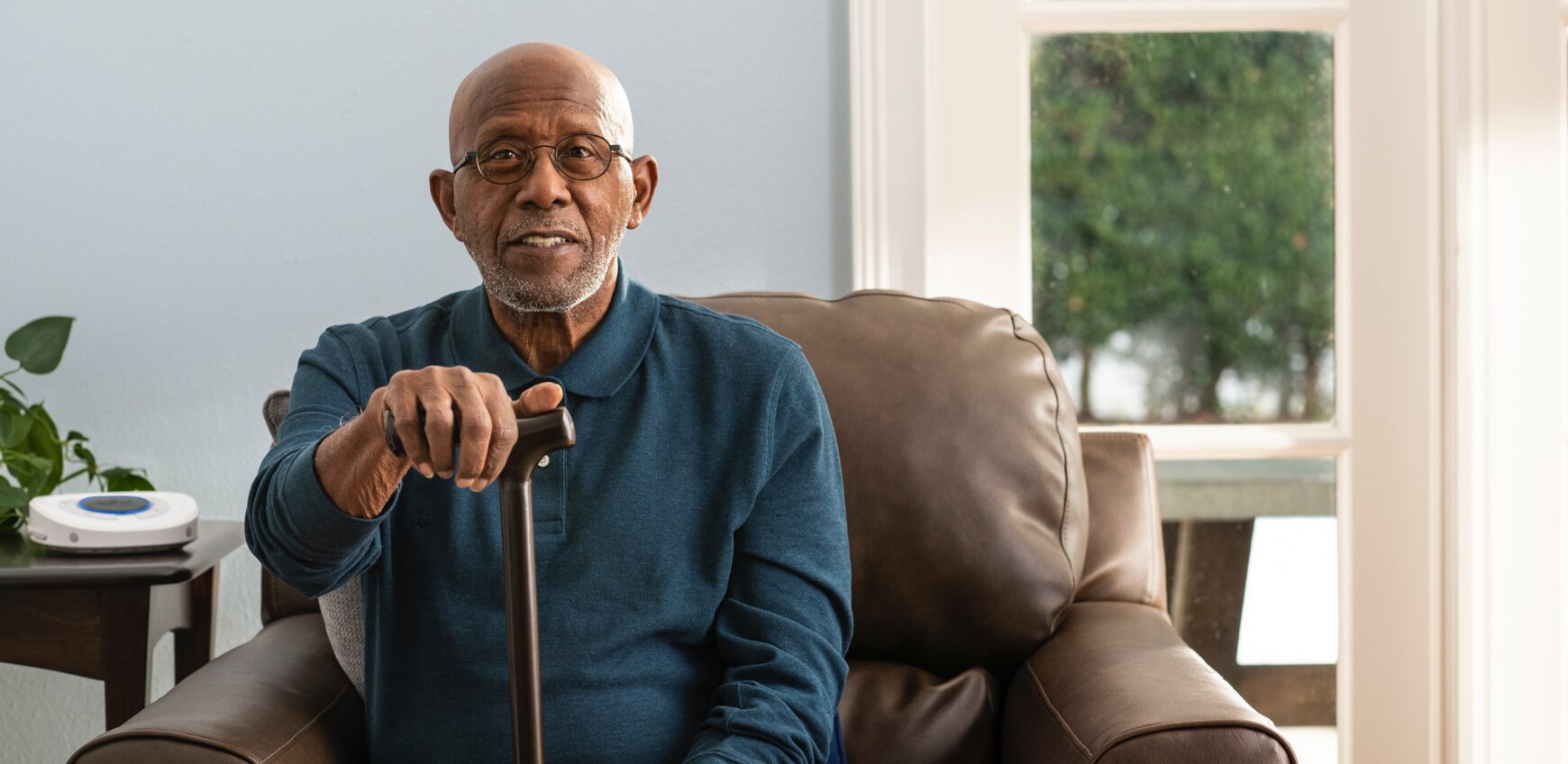 Smart Hub out of focus on living room side table. Elder gentleman sitting on couch holding walking cane.