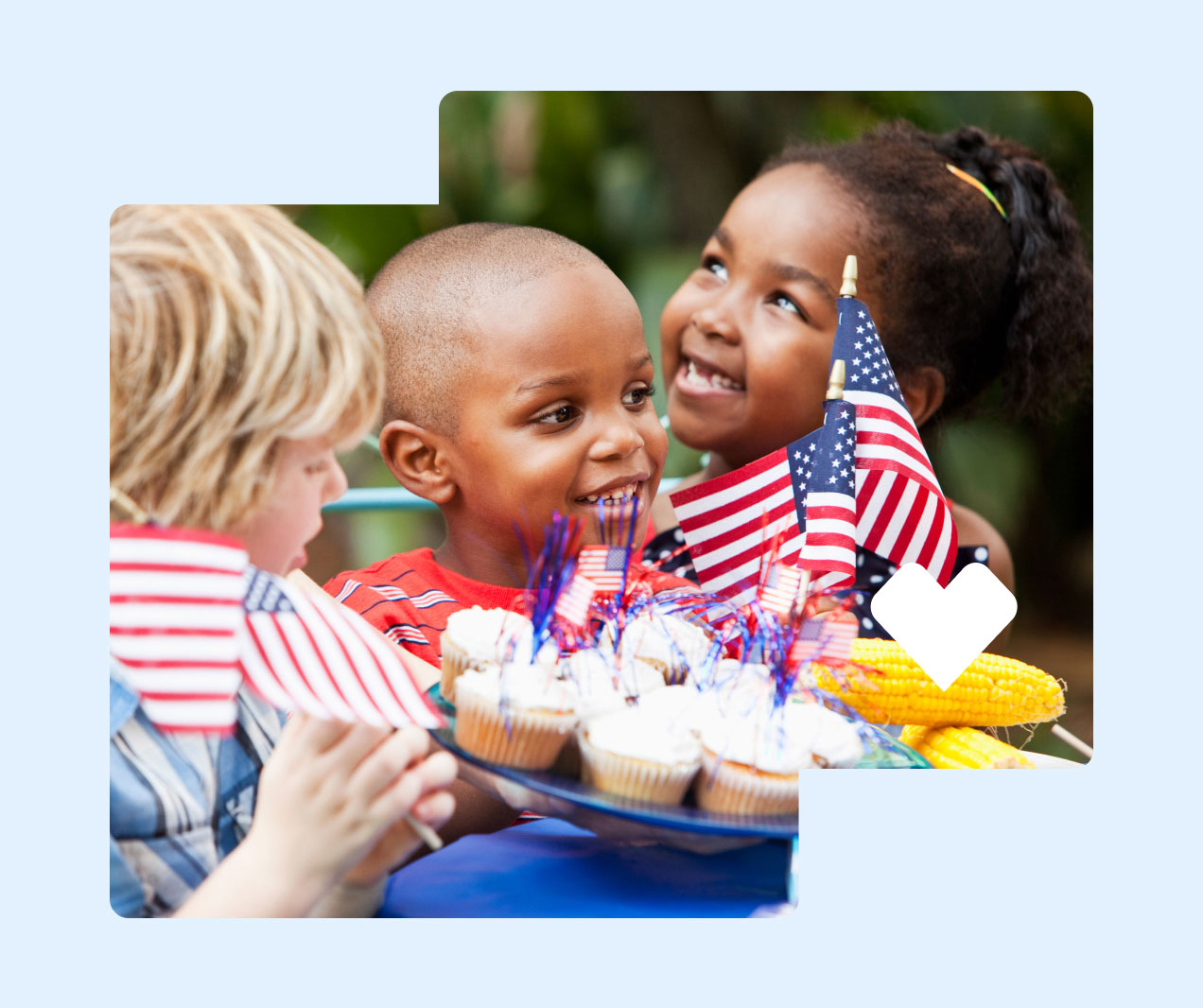 Children holding American flags sit at a table with cupcakes and grilled corn on the cob.