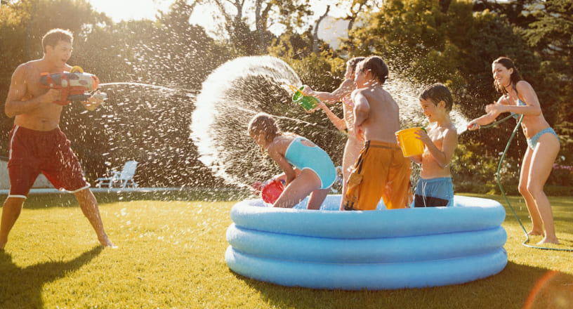 A parent sprays children in a kiddie pool with a garden hose while the children splash water back at the parent.