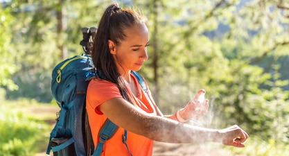 A person wearing a backpack on a hike stops to spray themselves with bug repellent.