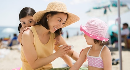 At the beach, a parent applies sunscreen to one of their young children while another young child peeks over the shoulder of the parent.