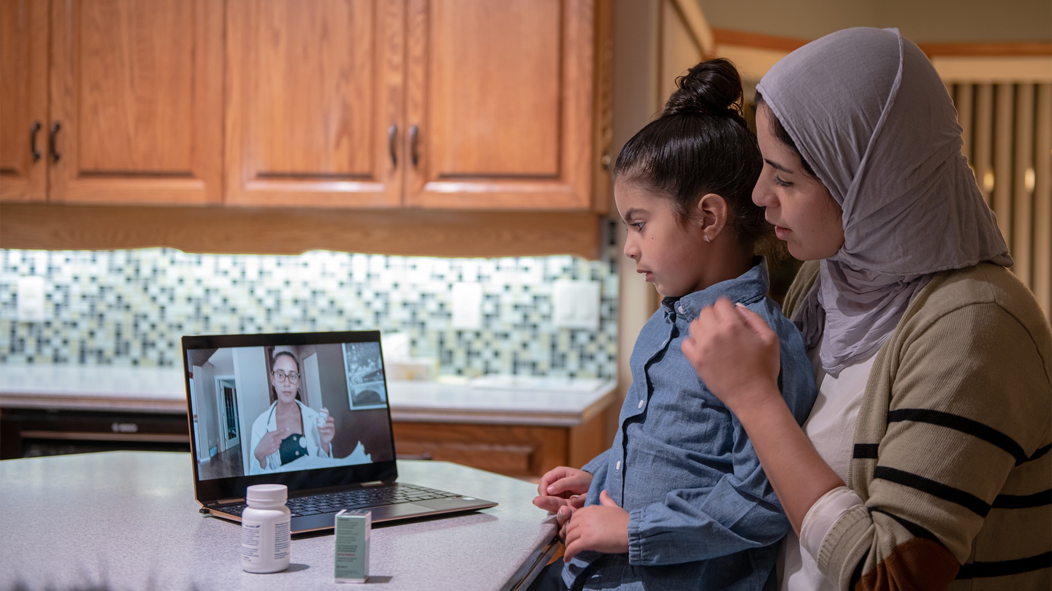Young mother in a hijab with her daughter on her lap speaking to a female doctor via video call on a laptop.