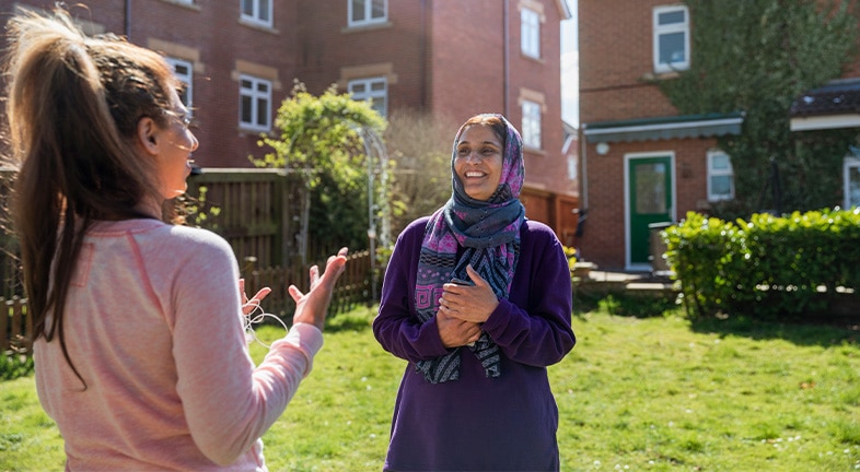 Mom wearing a hijab stands in her sunny backyard laughing at something her daughter said.