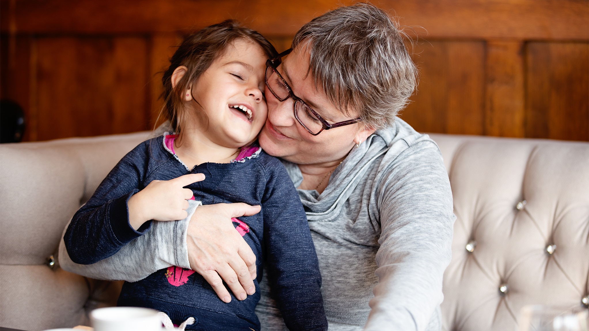 Mature woman with glasses embraces her granddaughter in a coffee shop.