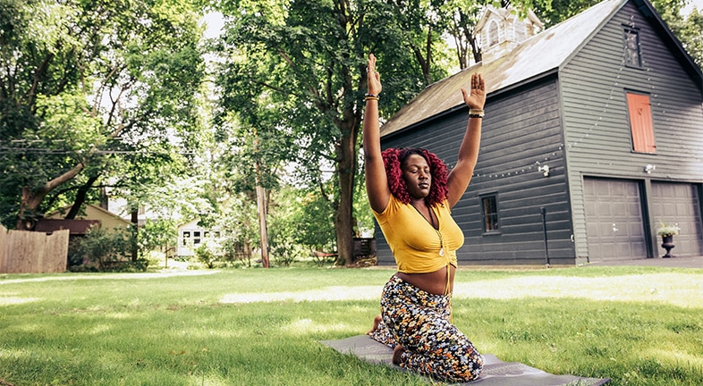 A young woman, in bright yellow top and floral yoga pants, kneels on an exercise mat outdoors in the grass among mature trees, in order to do a back stretch that involves reaching for the sky.