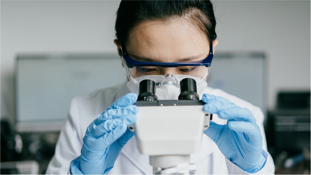 Un técnico de laboratorio lleva un traje de protección mientras mira por un microscopio de doble lente.