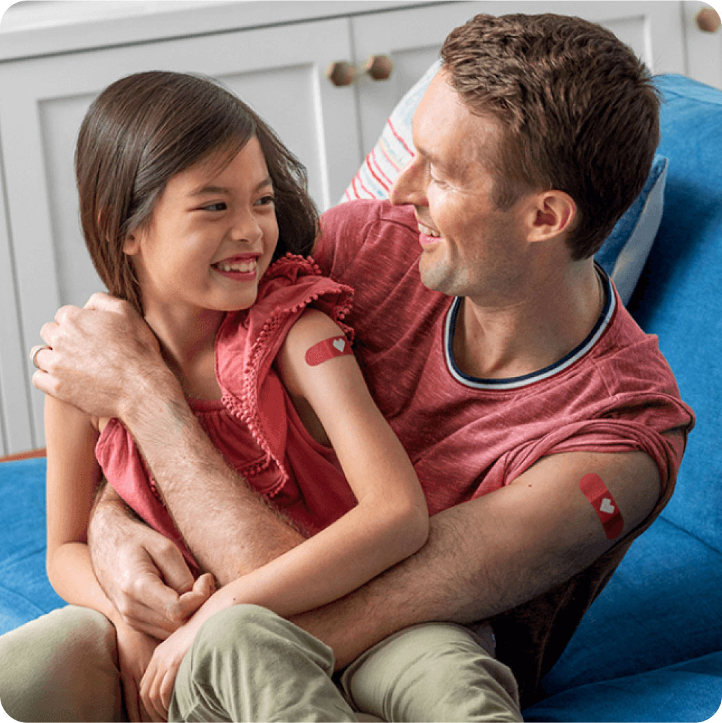 A smiling father and daughter compare arm bandages after their vaccinations.