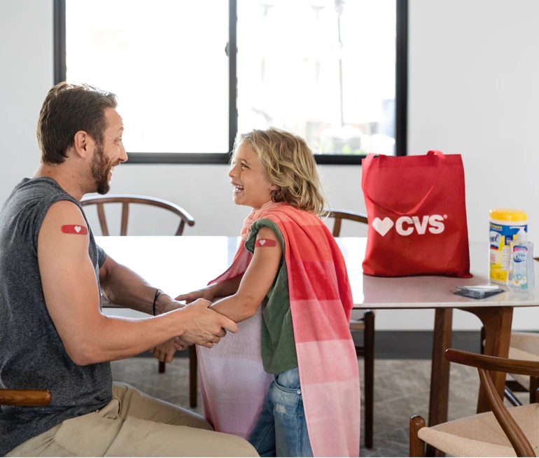 Imagen de hombre con venda en forma de corazón en el brazo y pequeña con capa. Hay una bolsa de productos CVS sobre la mesa.